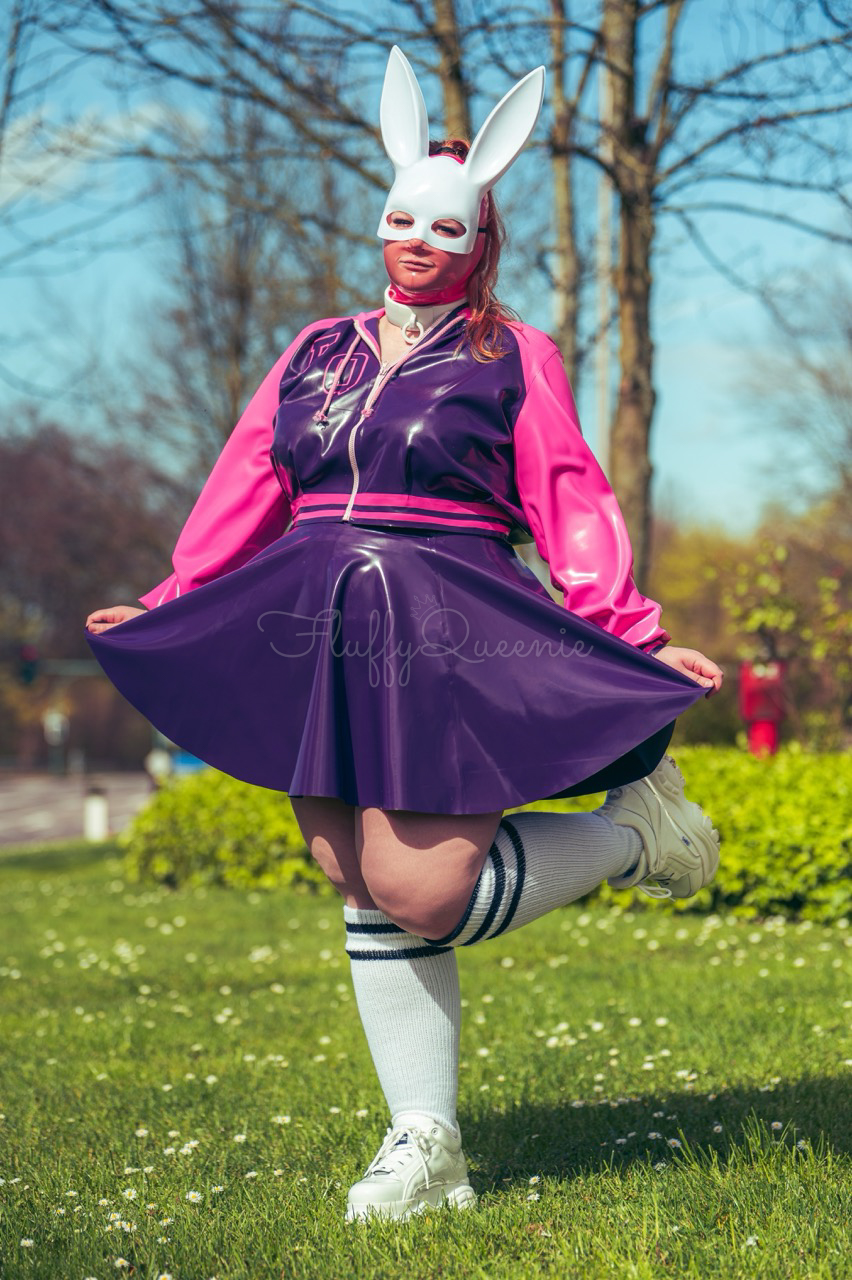 Plus-Size Latex Content Creator FluffyQueenie in a bunny outfit wearing white bunny mask, a white collar, purple and pink college jacket in cropped and a purple skater skirt. The knee socks are white and white buffalo shoes.