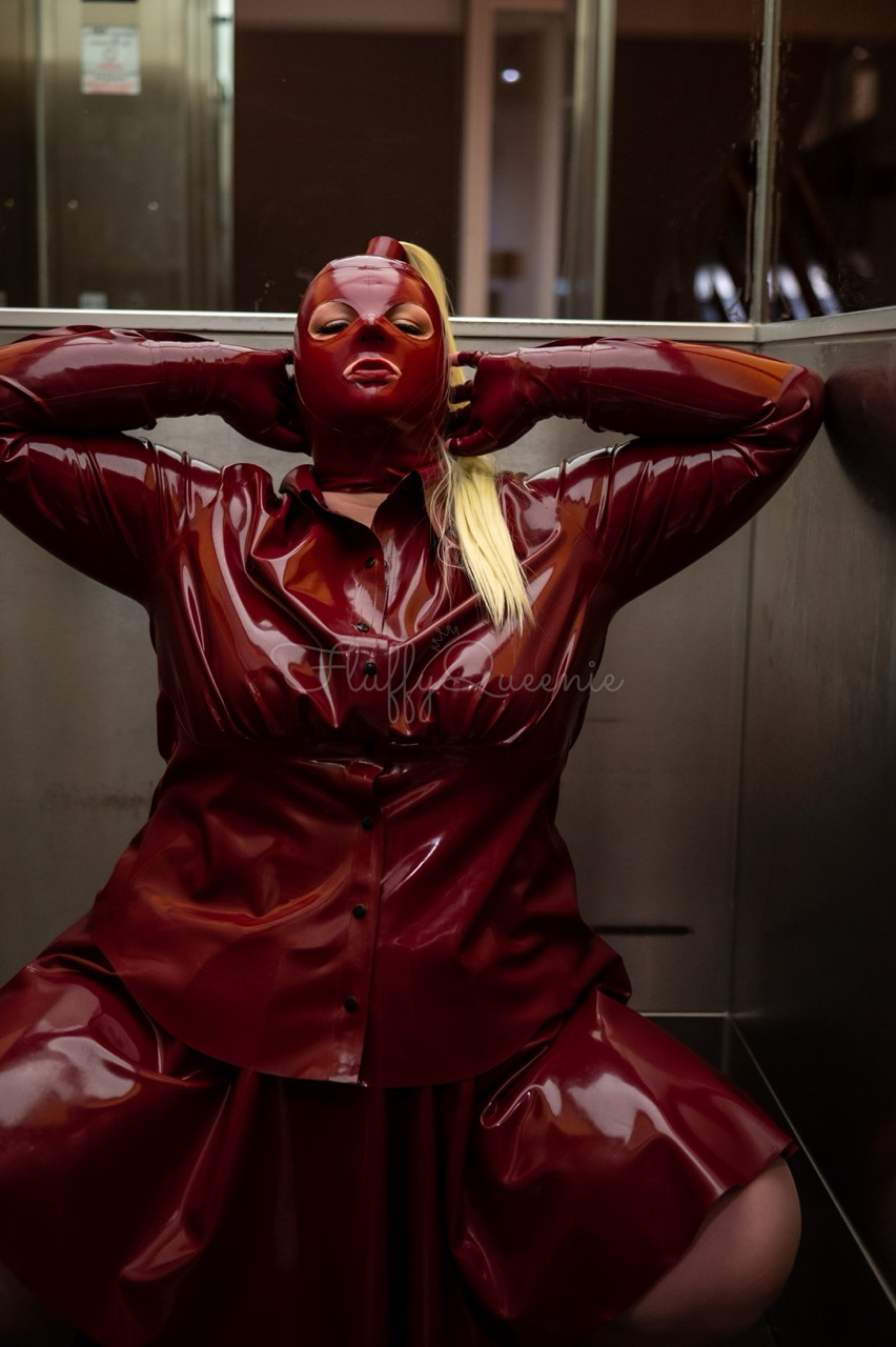Plus-Size Latex Model FluffyQueenie kneeling in a elevator wearing a plum latex mask, plum latex blouse and plum skater skirt.