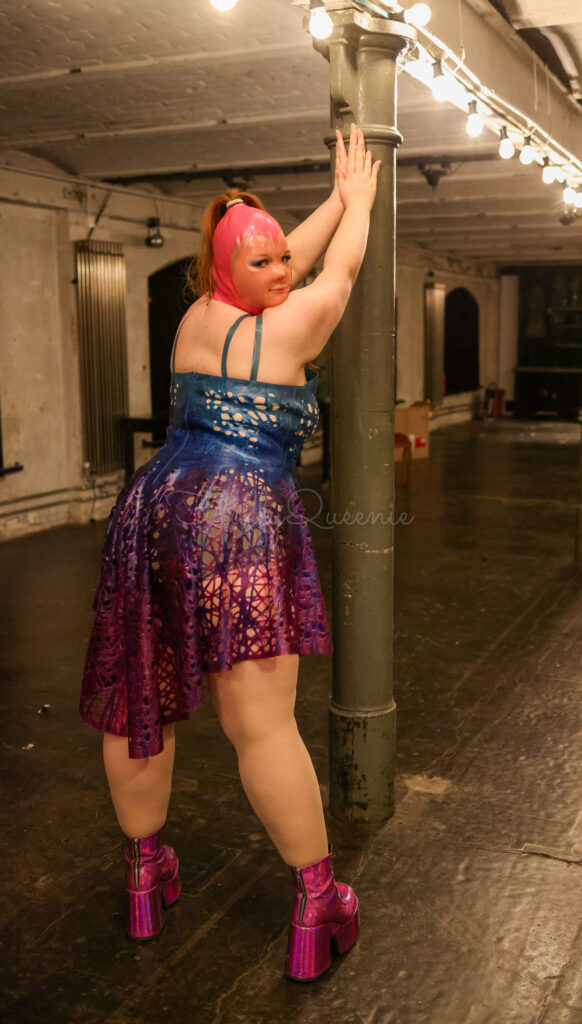 Plus-Size Girl posing in a lost place in a latex outfit. She wears a pink latex mask, and a blue-purple latex dress.