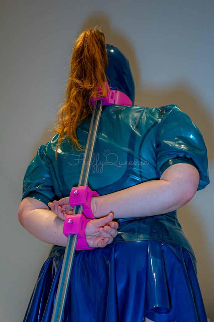Plus-Size Girl being tied up with a spreader bar behind her back. She wears a blue latex mask, transparent blue latex blouse and blue skater skirt.