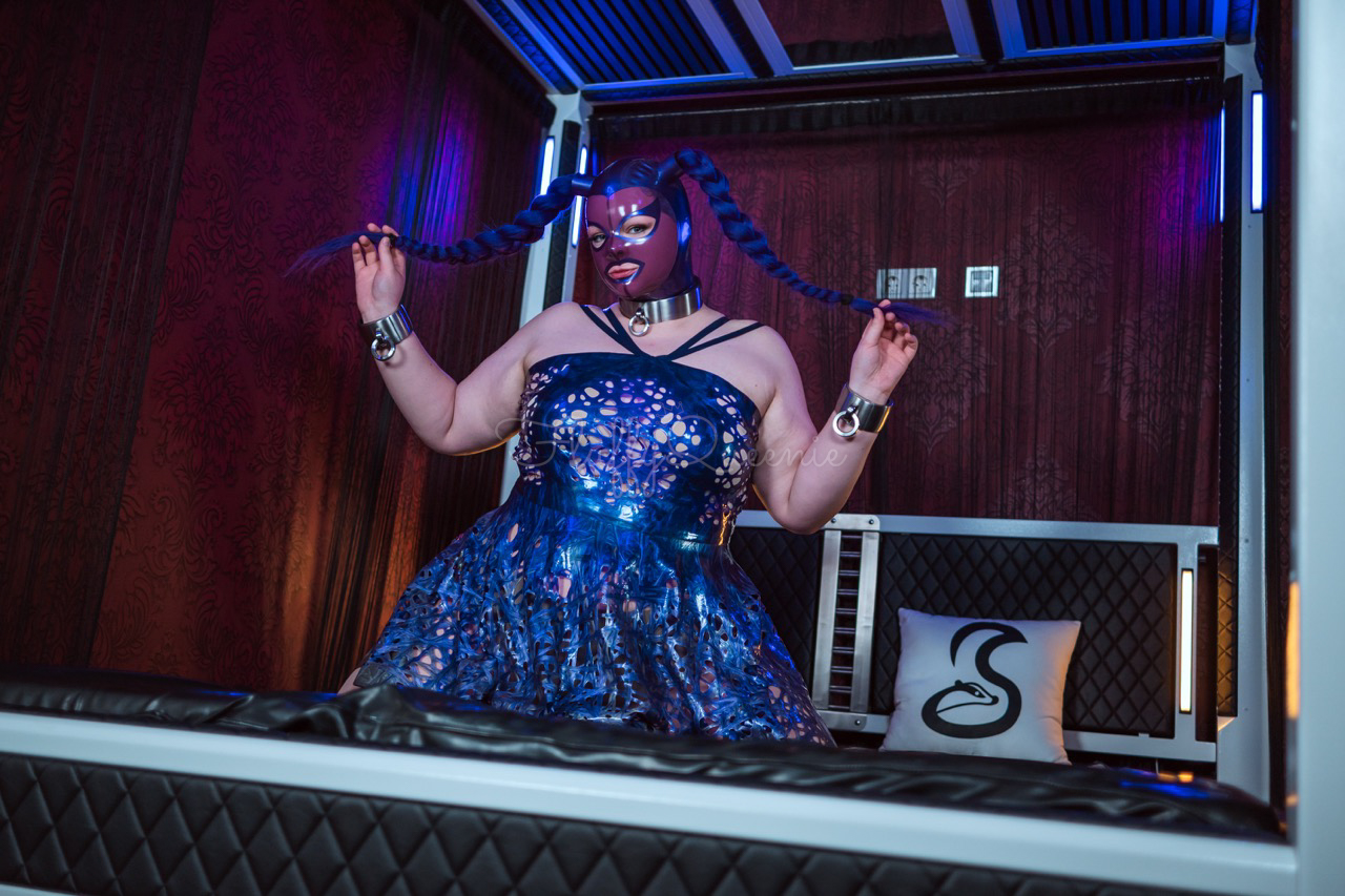 Plus-Size Girl kneeling on a skunkworx design bed, wearing a latex mask in purple with pigtales and a purple latex dress.